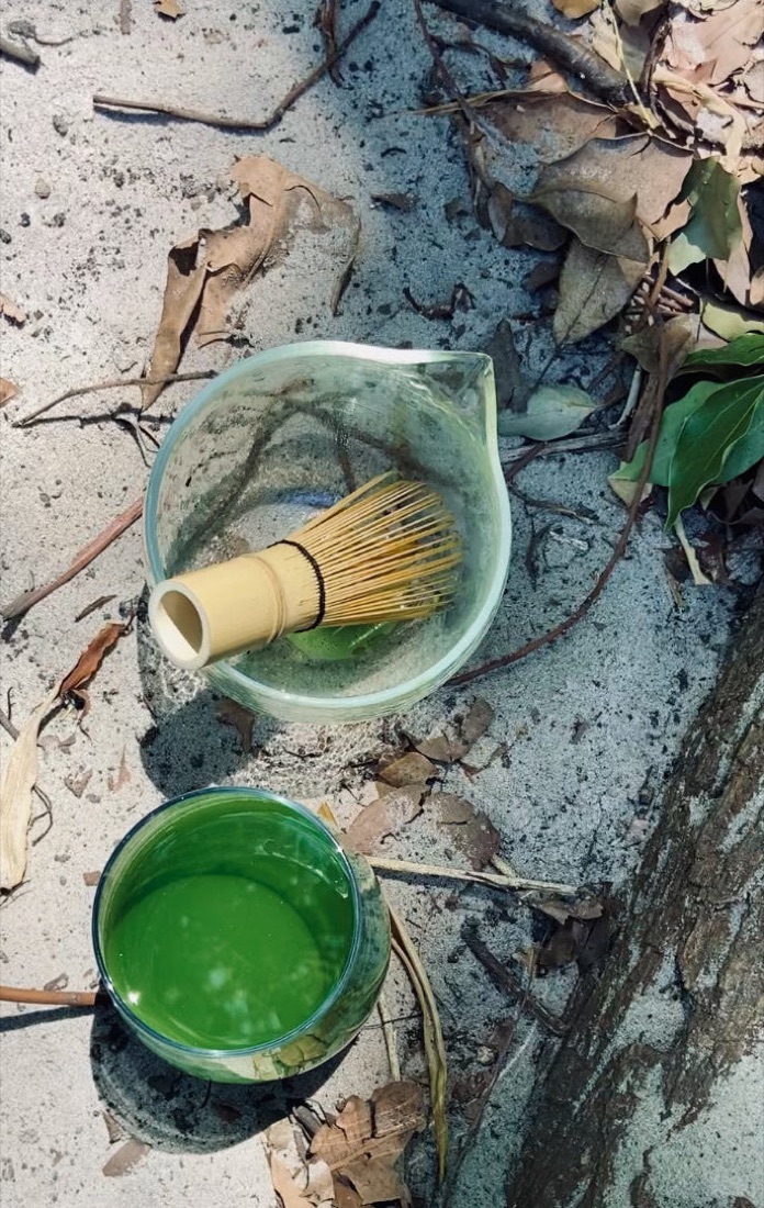 Glass matcha bowl with bamboo whisk and a cup of bright green matcha on sand among fallen leaves